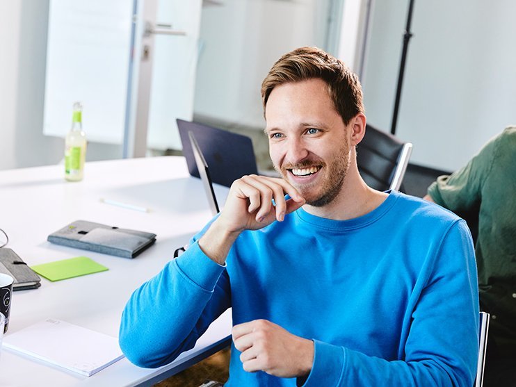 Ein lise-Entwickler in einem blauen Pulli sitzt an einem Tisch und guckt in die Luft