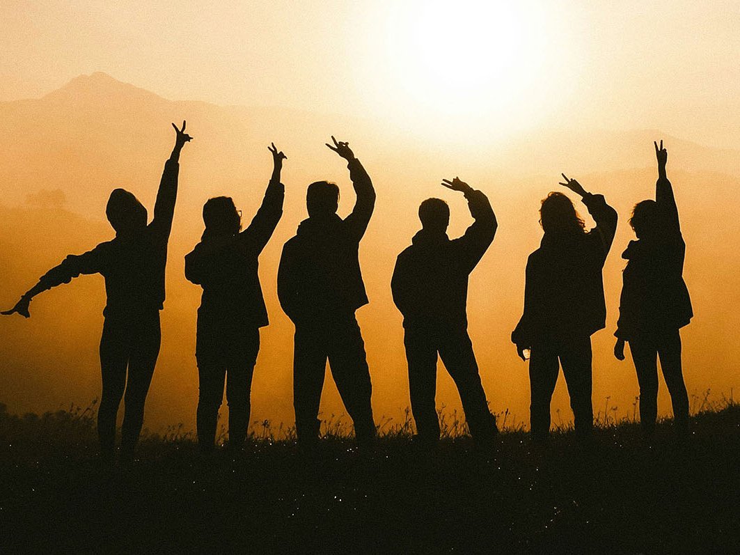 In einer Landschaft mit Sonnenuntergang sieht man die Schatten von 6 Menschen, die ihre Hände in die Luft halten. 