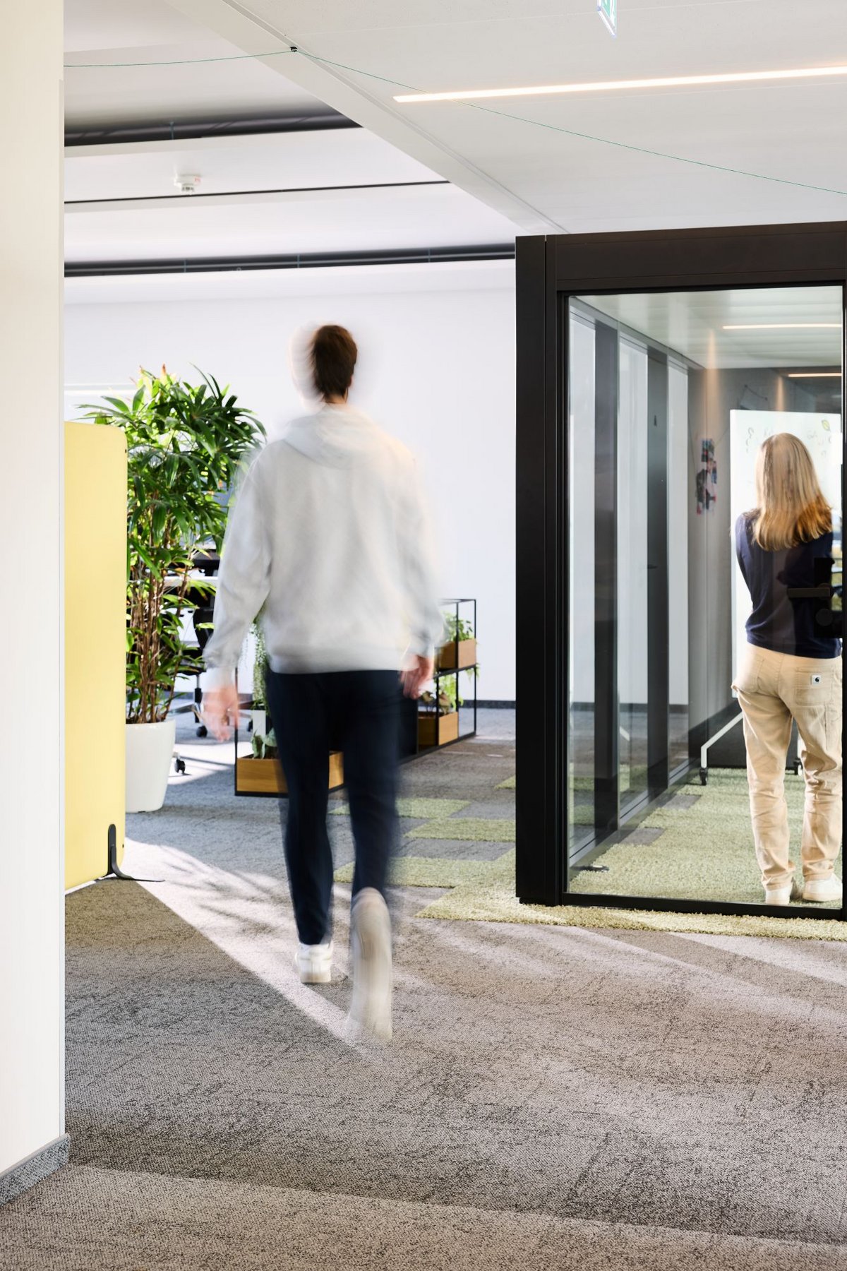 Eine lise-Entwicklerin hält ein Meeting in einem Gruppenraum währenddessen ein anderer Mitarbeiter an dem Raum vorbeiläuft