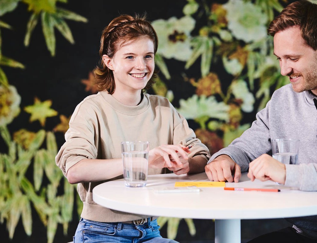 Ein Mädchen wird von einem lise-Mitarbeiter zur Fachinformatikerin ausgebildet, auf dem Tisch liegen dafür Zettel, Stifte und ein Glas mit Wasser