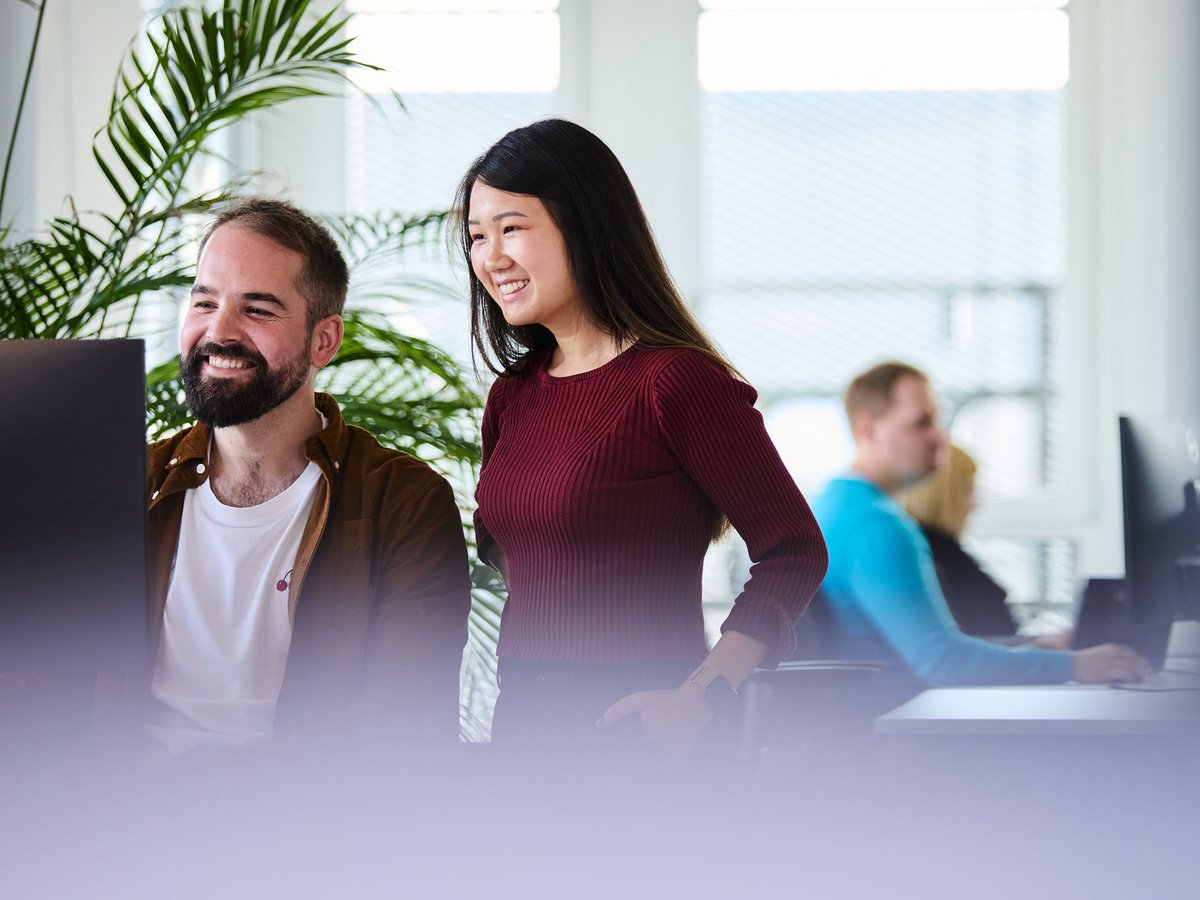 zwei lise-Entwickler*innen betrachten den Bildschirm des Computers vor ihnen, im Hintergrund steht eine große grüne Pflanze und man kann weitere lise-Mitarbeiter arbeiten sehen