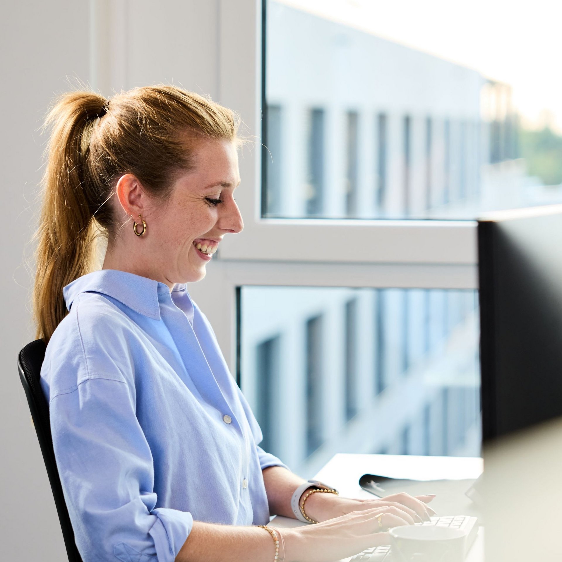 Eine lise-Entwicklerin mit einem hellblauen Hemd lächelt auf ihren Computer vor ihr