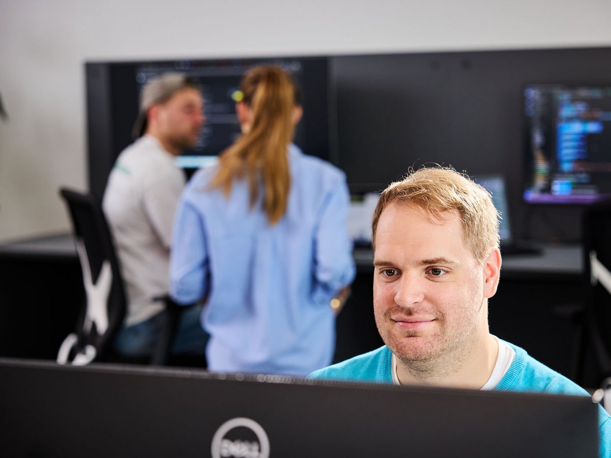 Ein lise-Entwickler ist im Vordergrund beschäftigt eine Software zu entwickeln, während sich zwei lise-Mitarbeiter*innen im Hintergrund austauschen