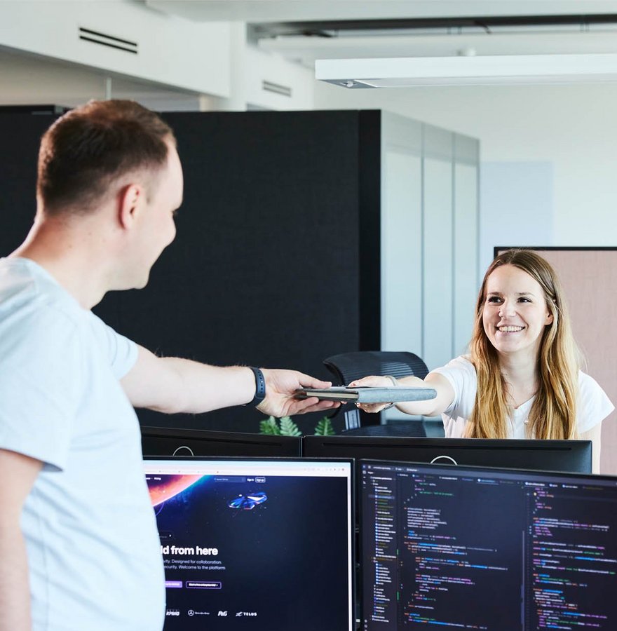 Zwei lise-Entwickler*innen stehen gegenüber voneinander und übergeben sich ein Notizbuch über Continuous Delivery