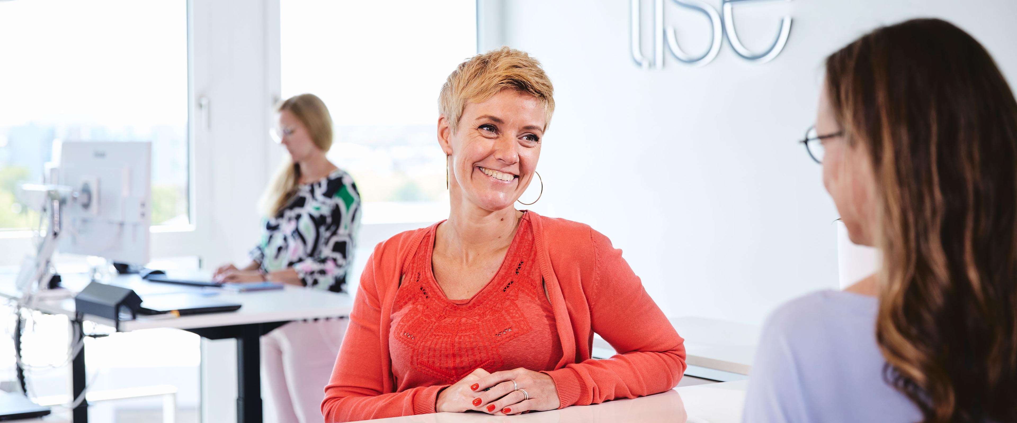 Zwei lise-Mitarbeiterinnen unterhalten sich lachend und im Hintergrund arbeitet eine weitere am Computer